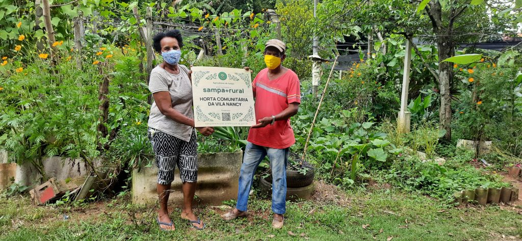 Decorative image from the São Paulo Project, two local people are holding a plaque. the plaque reads "sampa+rural. Horta comunitária da vila nancy." at the bottom of the plaque there is a QR-code.