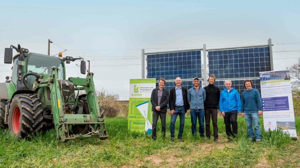 Decorative image of the project kick-off for the Agri-Photovoltaic Pilot Project. Six men are standing in front of some solar panels. There is a truck to the left. The men are bracketed by two posters with writing that is illegible in the photo.