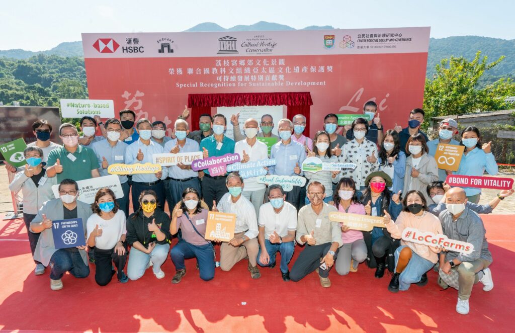 Decorative image depicting a group photo from the Hong Kong Project. Several of the people in the photo are holding signs depicting a SDG or a keyword. The SDGs depicted are 13, 12, 17, and 11. The keywords include: "Nature-based solutions", "Spirit of place", "Collaboration", "Rural Sustainability", "Climate Action", "LoCoFarms", "Urban-Rural Integration", "Bond for Vitality", and more.