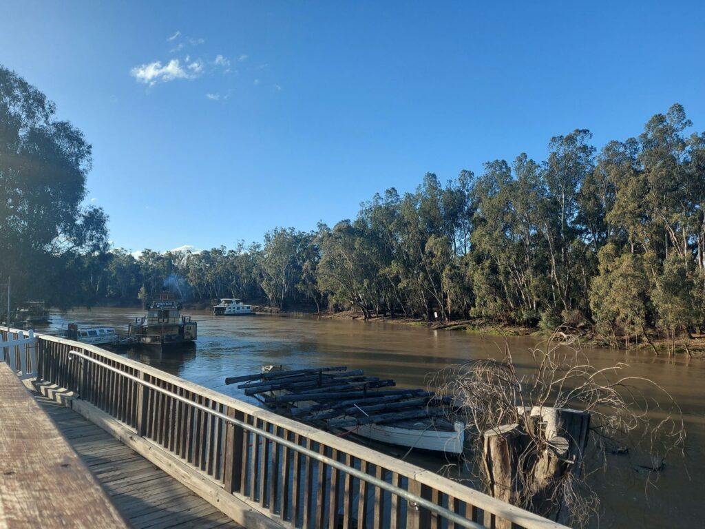 Image taken by Carolyn Hendriks and Christie Woodhouse. It shows a river from the view of a short, wooden bridge. There are five boats in the river. It is banked by trees. The sky is blue. The water is brown.