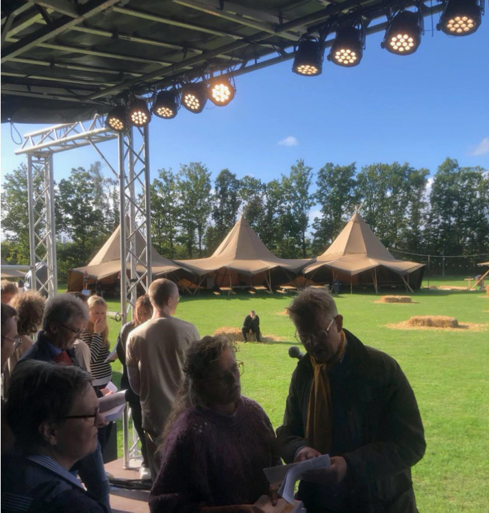 An image of the festival. The photo is taken on what might a stage where several people are mingling and discussing papers and books they are holding. There are three festival tents in the background and a field with hay piles to sit on.