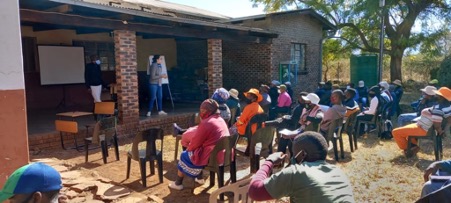 Image from the Dinkwayane Water Smart Project. Several people sitting in chairs outside are looking at a woman talking on a shaded porch.